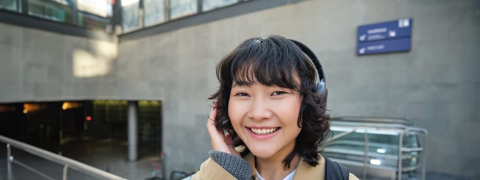 Close up of smiling brunette girl in headphones, listens music, travels around city, commutes to work, stands on a street.