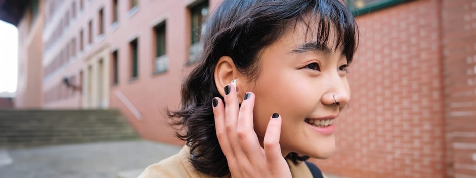 Close up portrait of young korean woman touches earphone, listens music in headphones with pleased smiling face, walks in city centre along the street.