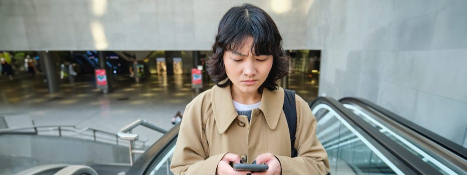 People in city. Portrait of girl looks concerned at smartphone screen with frowned worried face expression. Woman goes up escalator with mobile phone.