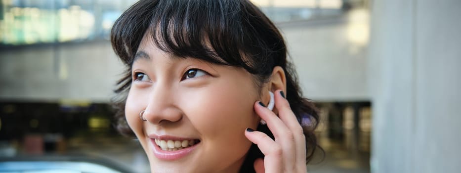 Close up portrait of smiling korean girl in headphones, listens music in wireless earphones, enjoys travelling around town with her favorite songs playlist.