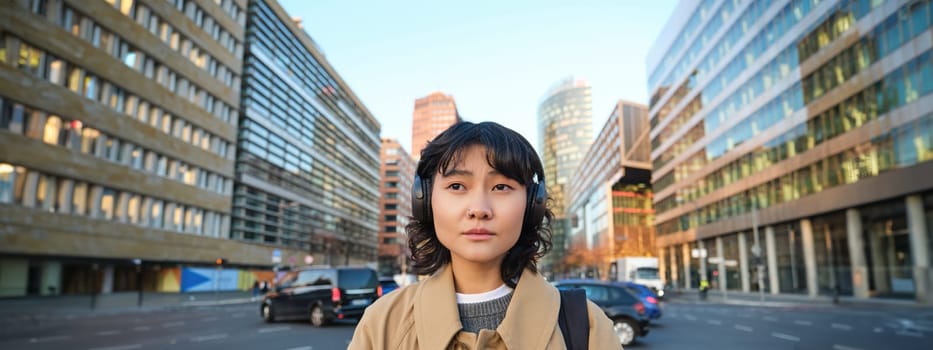 Portrait of concerned girl standing on street of city. Young korean woman in headphones, holding phone, looking worried and sad, reads bad news.