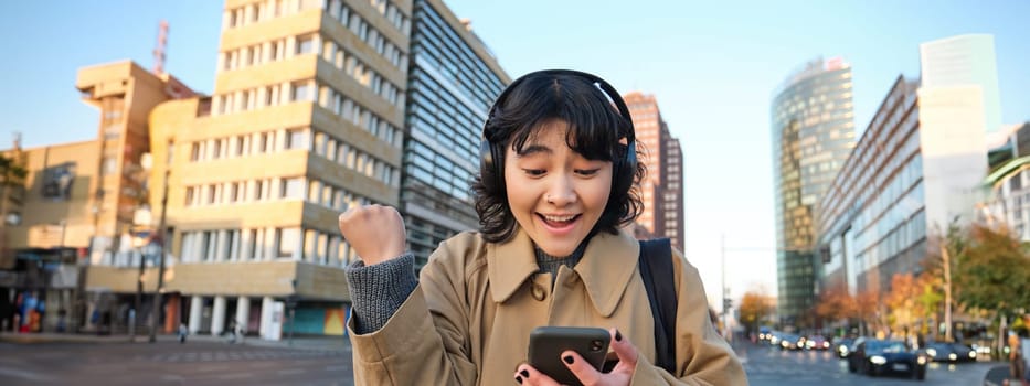 Young happy woman celebrating on street, holding smartphone and cheering, reacts amazed to good news, reads phone text message with surprised joyful face.