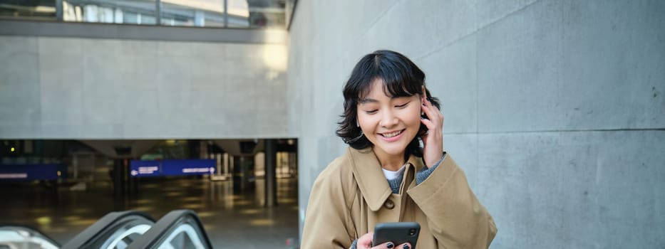 Young brunette woman commutes, goes somewhere in city, stands on escalator and uses mobile phone, holds smartphone and smiles, listens music in earphones.