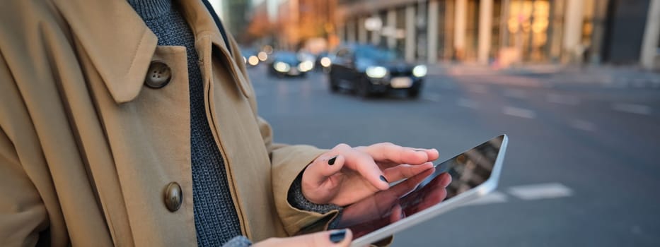 Close up photo of female hands holding digital tablet, girl looks at screen, checks application, stands on busy street filled with cars in afternoon.