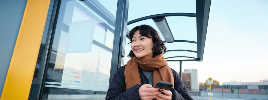 Cute smiling asian girl standing on bus stop, holding smartphone, wearing winter jacket and scarf. Woman commuting to work or university via public transport, stands on road.