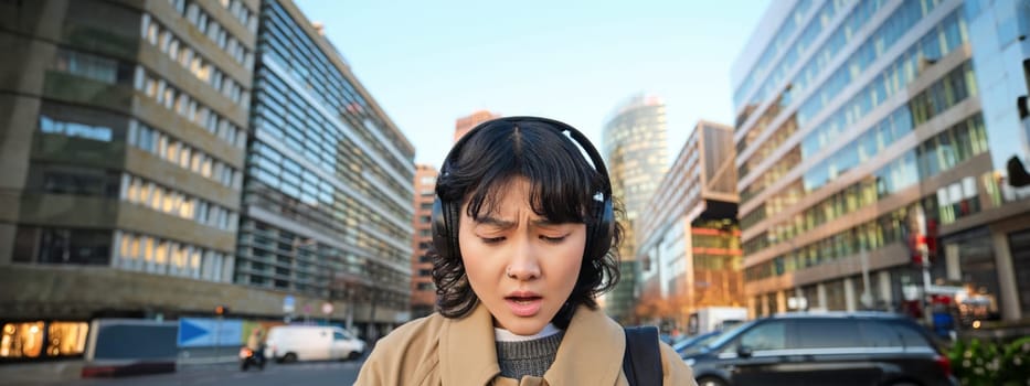 Portrait of young asian girl, student walks in city, listens music in headphones and uses mobile phone on streets, looks concerned at screen, reading bad news.