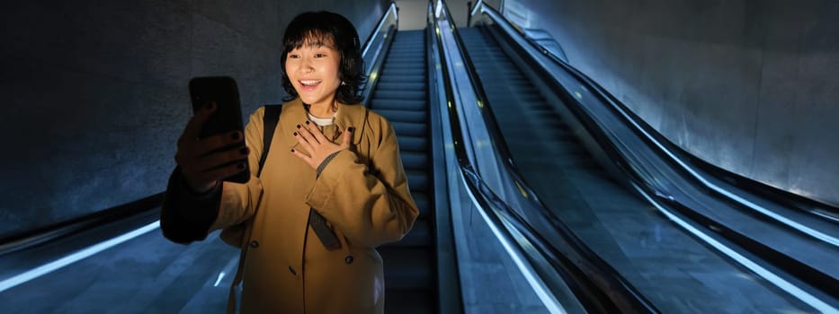 Surprised asian girl looks at smartphone screen, feeling amazed by smth she read on mobile phone, going down escalator in city, commuting to university.