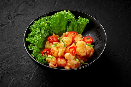 Appetizing wok fried basa fish pieces in spicy batter served in black bowl with fresh green lettuce and slices of red chili pepper. Asian style cuisine