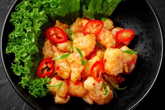 Pieces of deep fried white fish in crispy batter dressed with sweet chili sauce served with fresh lettuce, slices of spicy red pepper and scallions in black bowl, closeup view. Chinese style wok dish