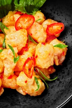 Closeup of wok fried spicy cod fillet pieces in fluffy batter sprinkled with chopped scallions and red chili pepper slices served in black bowl with greens. Asian style dish