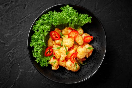 Delicious deep-fried shrimp in crispy beer batter with sweet chili sauce and spicy red pepper slices served in black bowl with fresh green lettuce and scallions