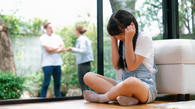 Stressed and unhappy young girl huddle in corner, cover her ears blocking sound of her parent arguing in background. Domestic violence at home and traumatic childhood depression. Panorama Synchronos