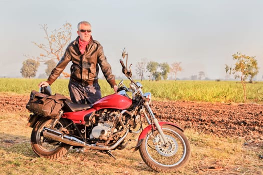 Gujarat, India, middle aged caucasian with red indian motorbike in a rural setting of seedings growing on a sugar cane farm