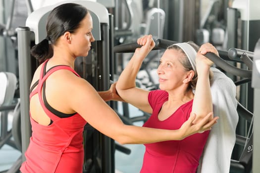 Personal trainer assist senior woman exercising on machine at gym