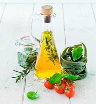 High angle view of fresh tomatoes and basil with uncooked green tagliatelle and a flask or carafe of golden olive oil on a white painted wooden surface.