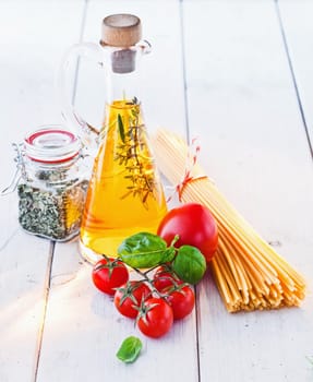 High angle view of fresh tomatoes and basil with uncooked spaghetti pasta and a flask of golden olive oil on a white painted wooden surface.