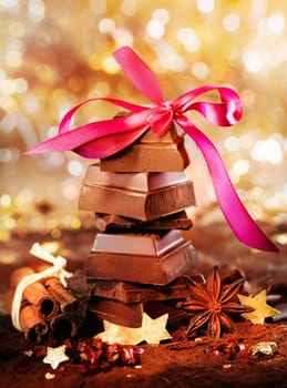 A stack of balanced chocolate pieces is surrounded by spices and stars and topped with a decorative pink bow against a bokeh of festive lights