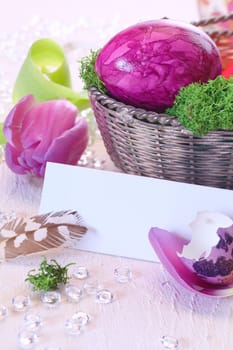 Easter still life centred around a blank white card with a pink marbled Easteregg in a basket alongside a tulip and broken eggshell