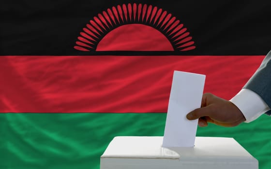 man putting ballot in a box during elections  in front of national flag of malawi