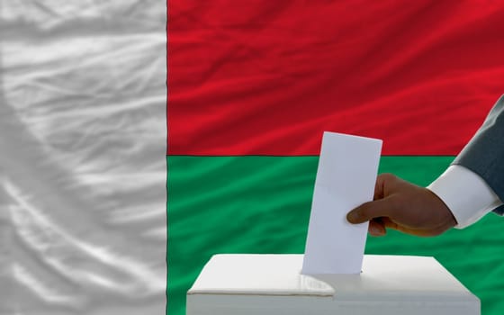 man putting ballot in a box during elections  in front of national flag of madagascar