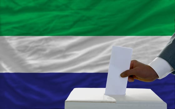 man putting ballot in a box during elections  in front of national flag of sierra leone