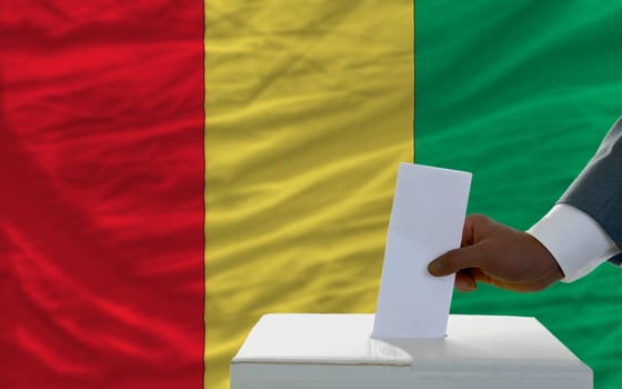 man putting ballot in a box during elections  in front of national flag of guinea