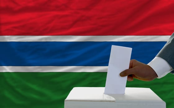 man putting ballot in a box during elections  in front of national flag of gambia