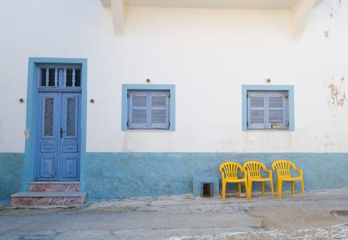 Santorini old white building with blue door and yellow chairs