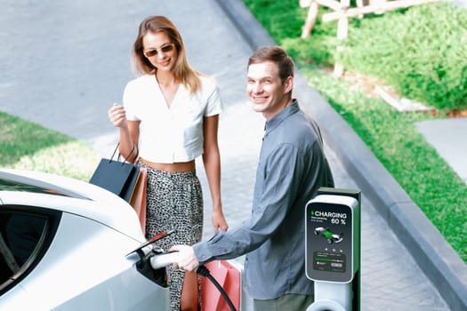 Young couple travel with EV electric car charging in green sustainable city outdoor garden in summer shows urban sustainability lifestyle by green clean rechargeable energy of electric vehicle innards