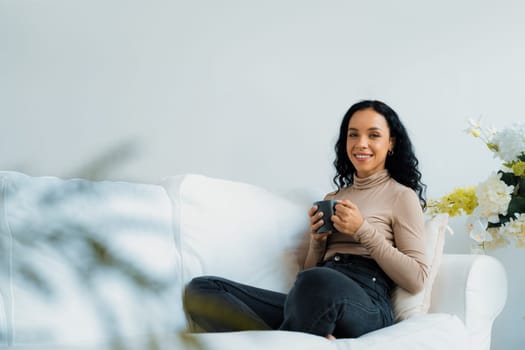 Happy woman drinking coffee on a sofa at home for crucial rest and relaxation. Portrait of young African American woman holding a cup.