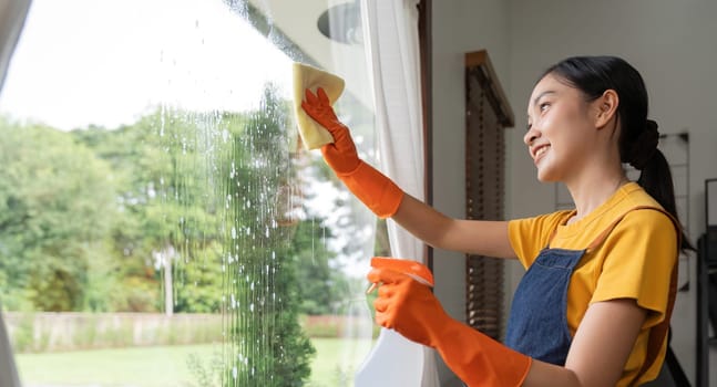 Young Asian housewife cleaning window glass with rags and detergent spray, cleaning house on the weekend.