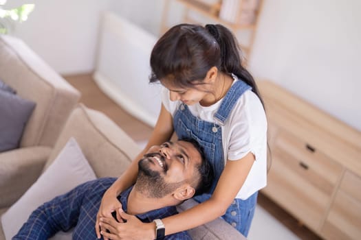 Happy lovely young Indian couple together at home, young wife hugging from behind her husband, sitting and rest on sofa in home, portrait of romantic multiracial couple in love.