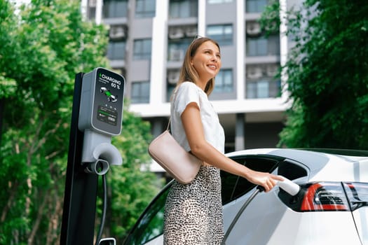 Young woman travel with EV electric car charging in green sustainable city outdoor garden in summer. Urban sustainability lifestyle by green clean rechargeable energy of electric BEV vehicle innards