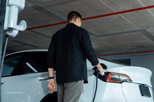 Young man travel with EV electric car to shopping center parking lot charging in downtown city showing urban sustainability lifestyle by green clean rechargeable energy of electric vehicle innards