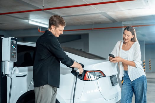 Young couple travel with EV electric car to shopping center parking lot charging in downtown city showing urban sustainability lifestyle by green clean rechargeable energy of electric vehicle innards