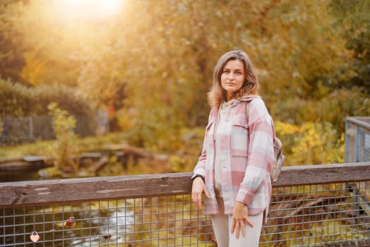 Portrait of cute young woman in casual wear in autumn, standing on bridge against background of an autumn Park and river. Pretty female walking in Park in golden fall. Copy space. smiling girl in the park standing on wooden bridge and looking at the camera in autumn season