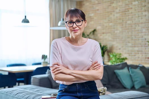 Portrait of positive confident middle aged woman at home. Smiling successful female with crossed arms looking at camera, living room interior background