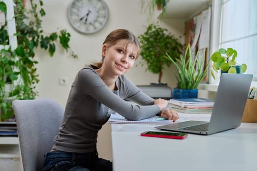 Portrait of teenage girl sitting at desk at home with laptop computer. Smiling 12, 13 year old young female student looking at camera. High school, adolescence, education, lifestyle concept