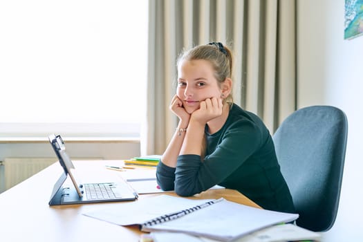 Portrait of smiling child girl sitting at desk at home with digital tablet textbooks, looking at camera. Education, knowledge, school, childhood concept