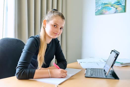 Portrait of smiling child girl sitting at desk at home with digital tablet textbooks, looking at camera. Education, knowledge, school, childhood concept