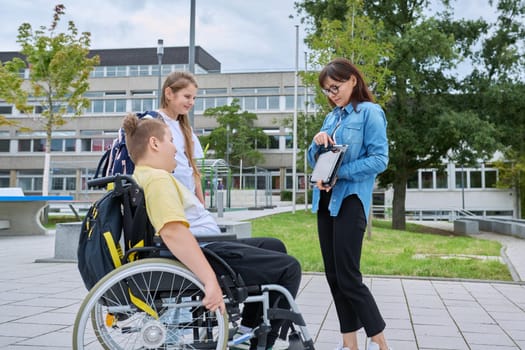 Female teacher with digital tablet and children classmates 10, 11 years old, boy in wheelchair and girl talking together outdoor, near school building. Education, disability, inclusiveness