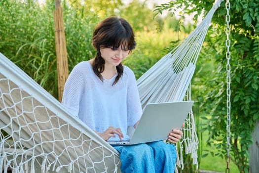 Teenage girl relaxing in hammock using laptop for leisure study. Adolescence, students, high school, technology, lifestyle, youth concept