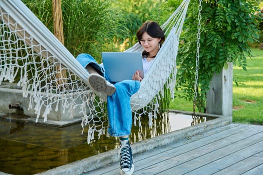 Teenage girl relaxing in hammock using laptop for leisure study. Adolescence, students, high school, technology, lifestyle, youth concept