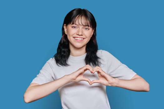 Smiling teenage female showing heart with hands looking at camera on blue color studio background. Young woman, university college student, showing her feelings emotion happiness romance