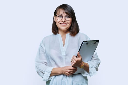 Confident middle aged business woman with clipboard looking at camera on white background. Mature smiling female teacher agent auditor psychologist supervisor posing with papers documents contracts