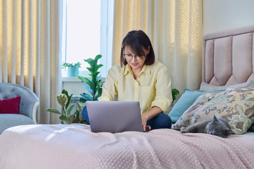 Middle aged woman at home on couch with laptop and cat. Mature 40s female looking at screen, on bed with pet. Work at home, technology, leisure, freelancing, lifestyle, people and animals concept