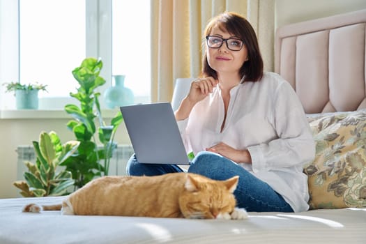 Middle aged woman at home on couch with laptop and cat. Mature 40s female looking at camera, on bed with pet. Work at home, technology, leisure, freelancing, lifestyle, people and animals concept