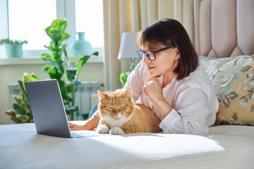 Middle aged woman at home on couch with laptop and cat. Mature 40s female looking at camera, on bed with pet. Work at home, technology, leisure, freelancing, lifestyle, people and animals concept