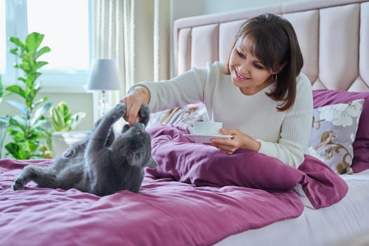 Middle-aged cheerful woman with cup of morning coffee in hand playing with cat in bed. Mature female owner touching pet, lifestyle friendship fun, people animals concept