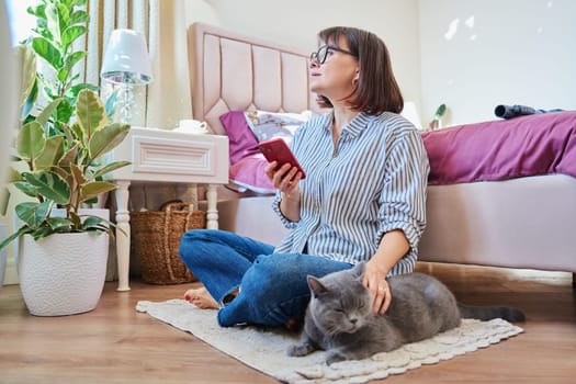 Home lifestyle, woman with cat, comfort calmness concept. Female sitting on floor on carpet using smartphone, pet gray british cat lying near owner